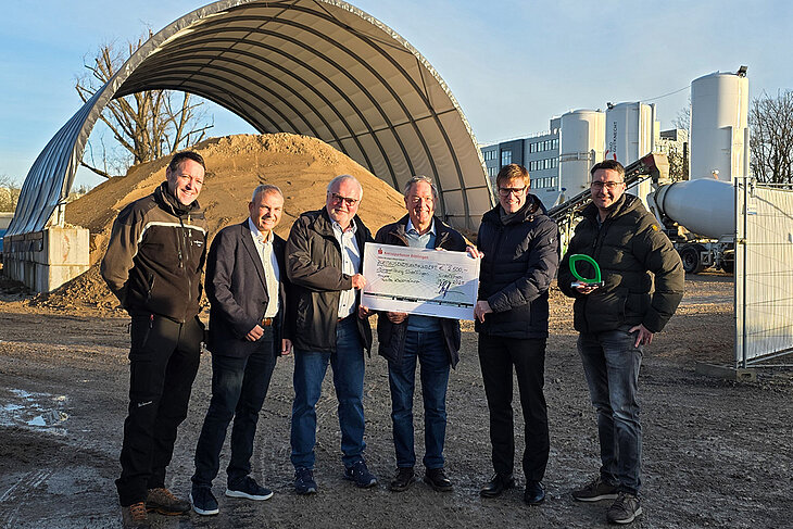 Auf dem Bild wird vom Geschäftsführer der Stadtwerke Sindelfingen ein Scheck über 2.500 Euro an die Bürgerstiftung und das Forstamt übergeben. Mit dabei ist auch OB Kleemann. Im Hintergrund sieht man die Mischanlage für Flüssigboden