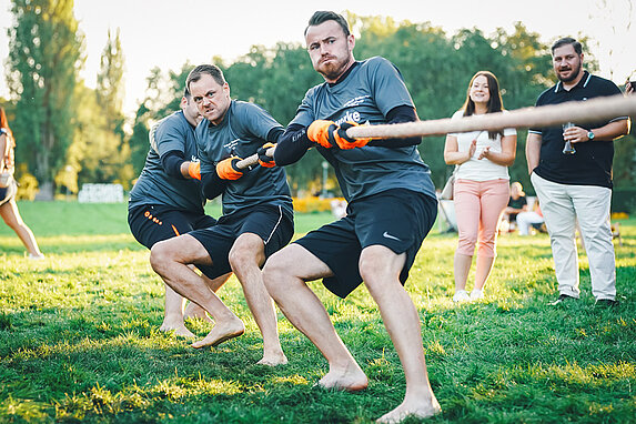 Drei Männer, allesamt barfuß und in Teamkleidung, beim Tauziehen auf einer Wiese. Sie beißen die Zähne zusammen und strengen sich sichtlich an. Im Hintergrund stehen weitere Personen und feuern sie an.