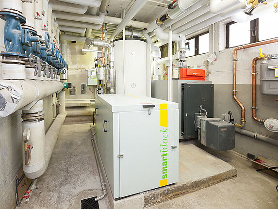 Mittig in diesem länglichen Technikraum steht ein weißes Blockheizkraftwerk für die Strom- und Wärmeerzeugung. Dahinter steht ein zylindrischer Pufferspeicher und ein silberner Gasheizkessel. An den Wänden und an der Decke sind viele Rohre und Leitungen verbaut.