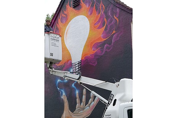 Das Graffiti zeigt eine große leuchtende Glühbirne, mit orange- und rosafarbene Blitzen. Darunter eine ausgestreckte Hand aus der blaue Blitze zur Glühbirne führen. Der Künstler steht auf einem Kran.