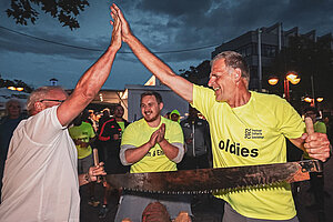Zwei Männer stehen sich an einem Holzsägebock gegenüber und geben sich ein High-Five. Die große Säge haben beide in der anderen Hand.