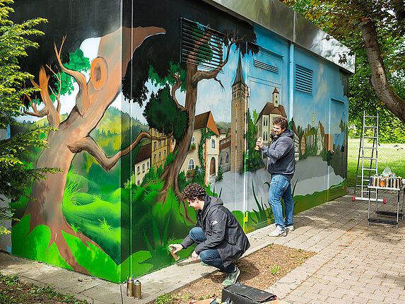 Ein technisches Gebäude mit einem grossflächigen Graffiti. Die zwei Künstler Stehen bzw. hocken um das Bild zu sprühen. Einige Flächen sind noch nicht bearbeitet.