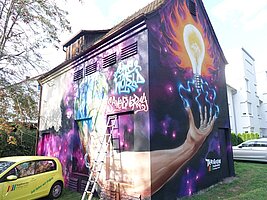 Ein Haus mit einem großflächigen Graffiti. Motiv des Graffitis: Aus der Erdkugel wächst ein Arm heraus. Die Finger sind nach oben gespreizt und blaue Blitze führen zu einer Glühbirne. Diese leuchtet hellgelb, drumherum sind orangefarbene Flammen. Die Worte "Save the World" und "Save Energy" stehen zusätzlich neben der Erdkugel.