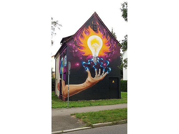 Ein Haus mit einem großflächigen Graffiti. Motiv des Graffitis: Eine Hand deren Finger nach oben gespreizt sind und blaue Blitze führen zu einer Glühbirne. Diese leuchtet hellgelb, drumherum sind orangefarbene Flammen.
