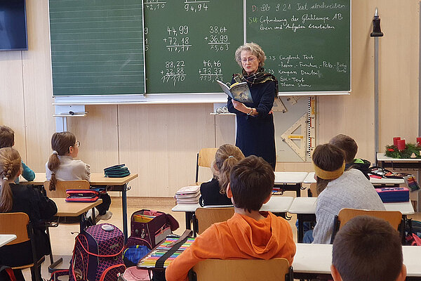 Eine Lehrerin steht vor der Tafel und liest aus einem Buch. Die Schulkinder sitzen alle auf ihren Plätzen und hören aufmerksam zu.