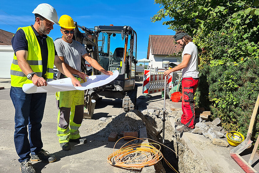 Zwei Bauarbeiter mit Schutzhelmen betrachten den Bauplan. Ein dritter Arbeiter vermisst einen Graben.