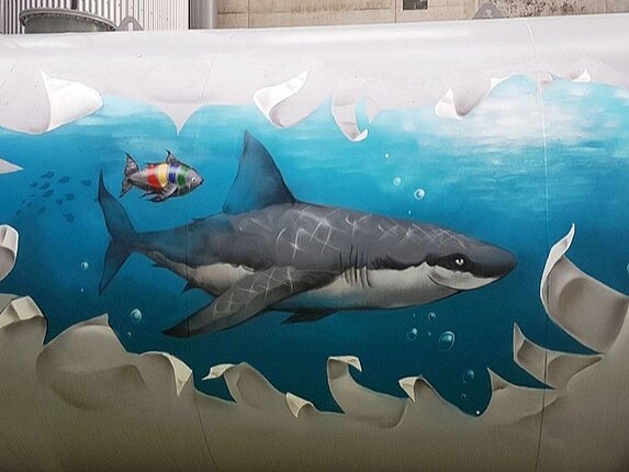 Ein Graffiti auf einem Öltank. Das Motiv: Es scheint als wäre da ein riesiger Riss im Tank, die Ränder sind ausgefranst und mann kann ins Ineere schauen. Dort schwimmt ein grinsender Hai und ein kleiner Fisch in den Farben der Stadtwerke Sindelfingen in hellblauem Wasser. 