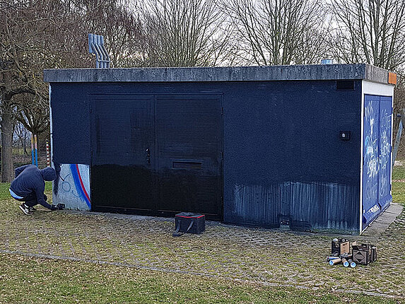 Ein kleines technisches Gebäude hat die erste Grundfarbe (ein dunkles blau, fast schwarz) bekommen. Der Künstler hockt an einer Seite des Gebäudes und besprüht es gerade.