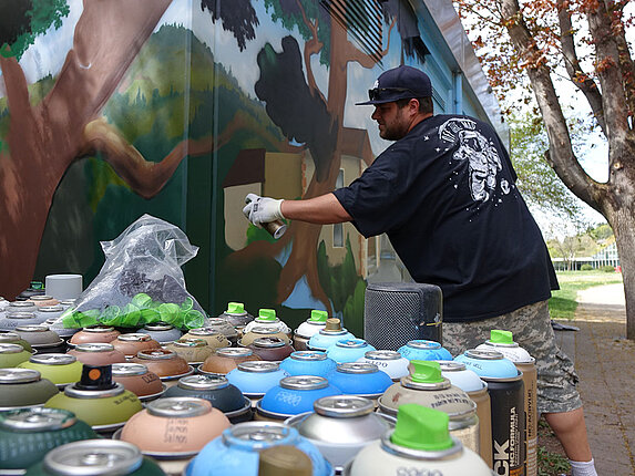Der Graffiti-Künstler sprüht gerade ein Graffiti auf eine Hauswand. Im Vordergrund stehen dutzende Spraydosen in unterschiedlichsten Farben.