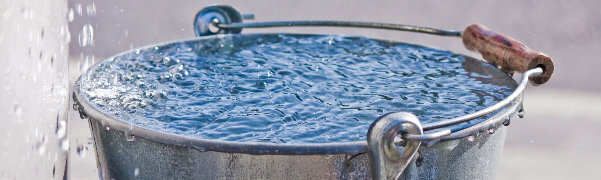 Ein metallener Wassereimer mit Holzgriff ist bis zum Rand mit Wasser gefüllt. Nachlaufendes Wasser läuft langsam über den Rand.