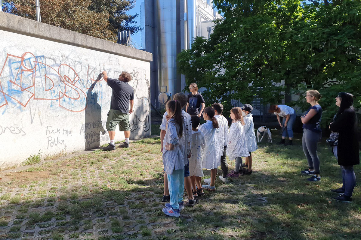 Ein Graffiti-Künstler sprüht erste Umrisse des neuen Motivs an eine Hauswand. Jüngere Kinder stehen davor, schauen zu und warten, um mitmachen zu können.