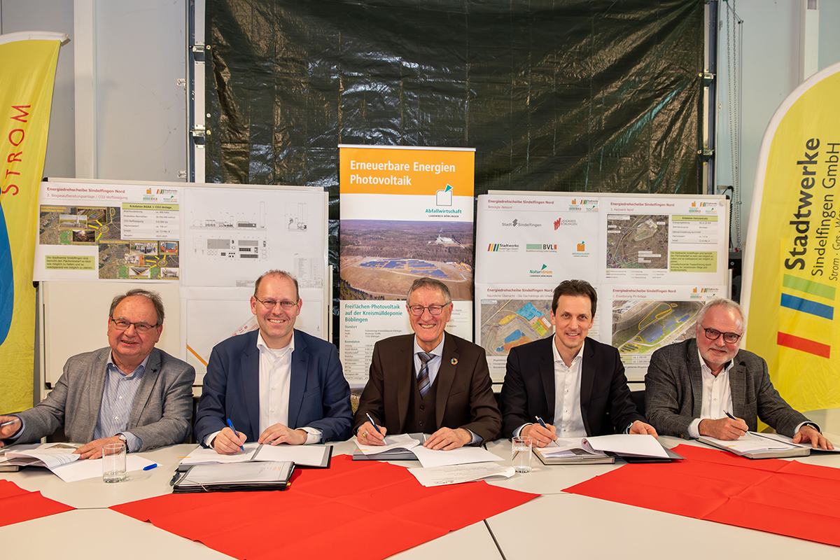 Energiedrehscheibe Nord Die beteiligten Personen sitzen nebeneinander an einem Tisch und unterzeichnen einen vertrag. Hinter ihnen sind Informationstafeln für die neue Fotovoltaikanlage aufgestellt.