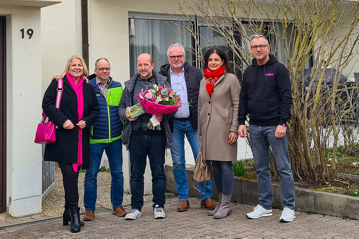 Gruppenbild von Herrn Hoffmann und anderer Personen vor einem Einfamilienhaus. Ein Mann hält einen Blumenstrauß in den Händen.