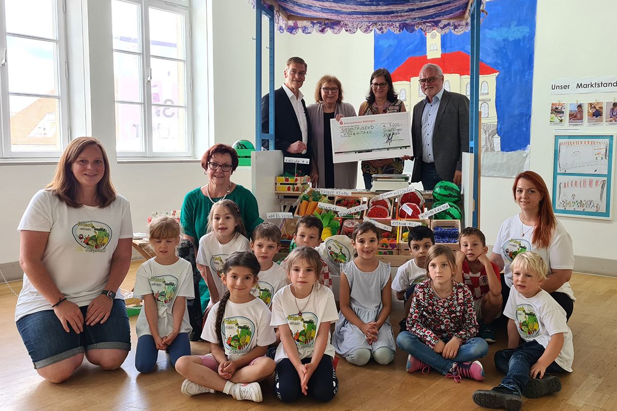 Stadtwerke unterstützen kidsatkita Symbolische Scheckübergabe in einem Kindergarten. Die beteiligten Personen stehen hinter einem kreativen Marktstand, die Kindergartengruppe sitzt davor.